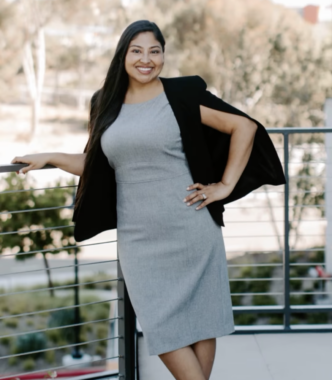 A professional photo depicts a young woman is a knee-length gray dress with a black shawl. She has long, dark hair that's gathered over her right shoulder, and she's lining on a railing of what appears to be a balcony. She's maybe two or three stories high, and a road and trees are visible behind her.